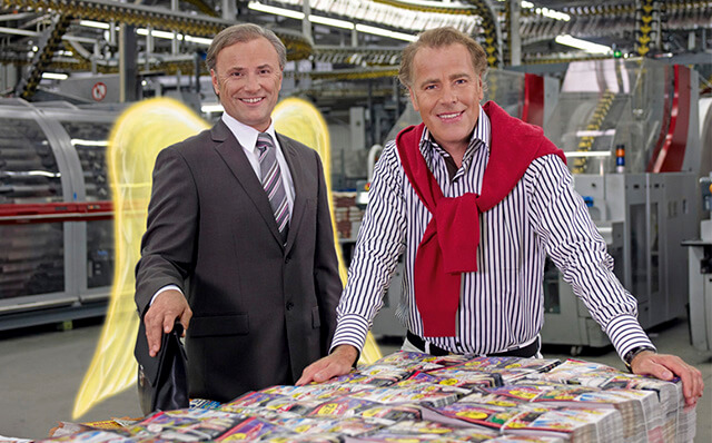 Ein Schutzengel im Anzug und eine Druckeri-Mitarbeiter stehen vor einem Stapel mit Druckerzeugnissen.