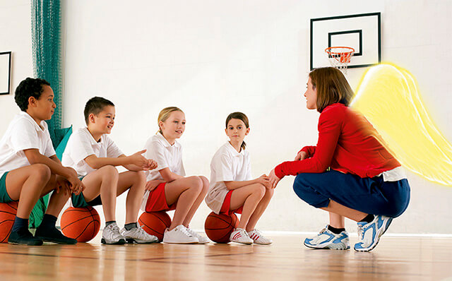Eine Trainerin mit Schutzengelflügeln kniet vor 4 Kindern, die auf Basketbällen sitzen.