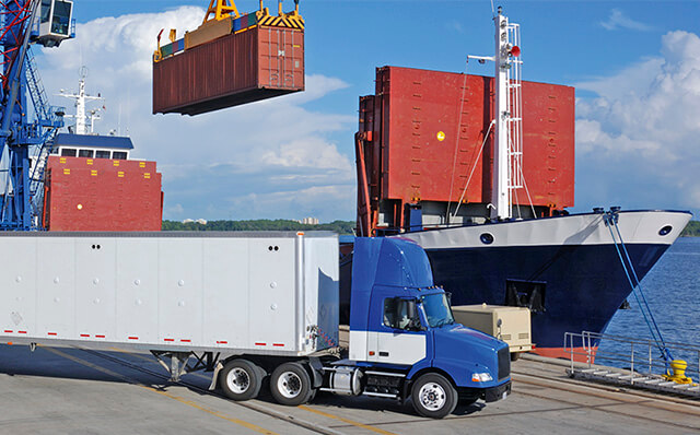 Im Vordergrund des Bildes steht ein großer LKW, dahinter belädt ein Kran ein Schiff mit einem Container.