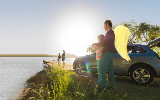 Ein Vater mit Schutzengel-Flügeln steht mit seinem Kind am See. Im Hintergrund das parkende Auto.