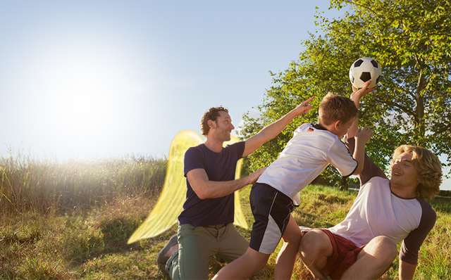 Private Unfallversicherung: Ein Vater spiel mit seinen Kindern Fußball. Der Vater hat Schutzengel-Flügel.